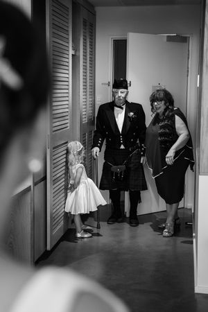 Black and white wedding photo showing people in formal attire including a man in a kilt greeting a young flower girl.