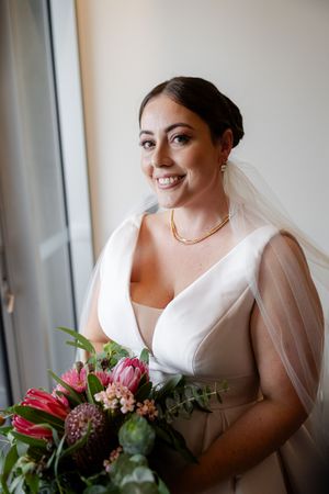 A bride in a white wedding dress holds a vibrant bouquet of pink tulips and greenery while standing by a window.