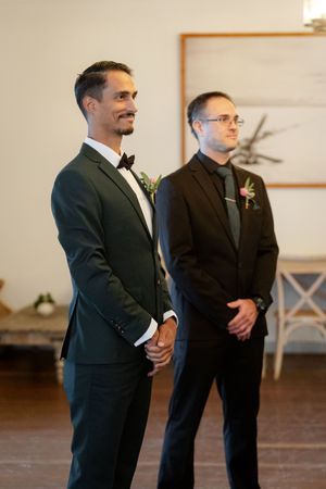 Two groomsmen stand side by side at a wedding ceremony, wearing formal black suits and boutonnieres.