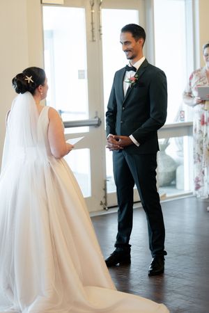 Wedding couple exchanges vows in a bright sunlit venue with white columns and elegant decor.