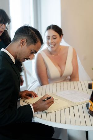 A pair signs documents at a light colored wooden table during what appears to be a wedding ceremony.