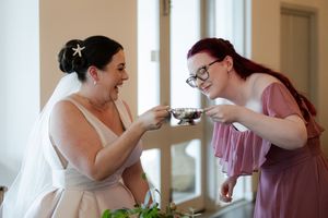 Bridesmaids in mauve dresses help with wedding preparations in a sunlit room.