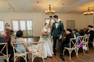 Wedding couple walking down the aisle in an elegant venue with cross-back chairs and chandeliers.
