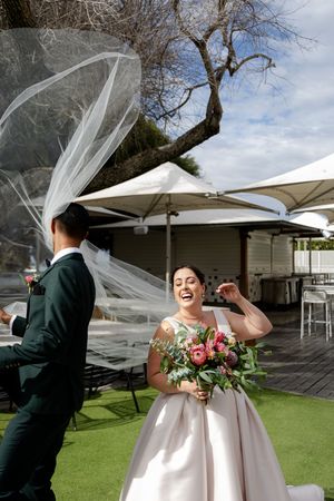 A joyful wedding moment captured outdoors under white umbrellas with flowing veil and pink bouquet.