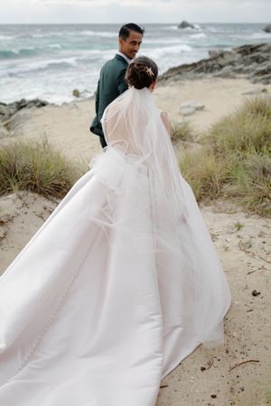 A couple in wedding attire walks along a rocky beach with ocean waves and grassy dunes in the background.