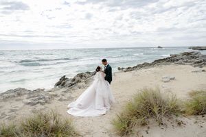 A romantic coastal wedding photo showing a couple on sandy dunes overlooking the ocean.