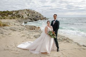 A couple in wedding attire stands on a rocky beach coastline with turquoise waters and dramatic cliffs in the background.