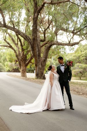 A bride in a flowing white gown and groom in a black tuxedo stand together on a tree-lined country road.