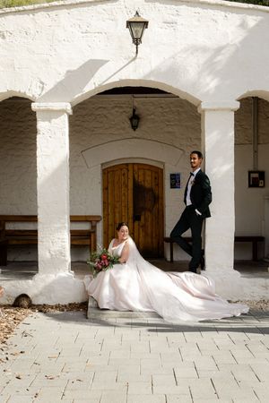 A wedding couple poses elegantly beneath white arches and lanterns at a historic building entrance.