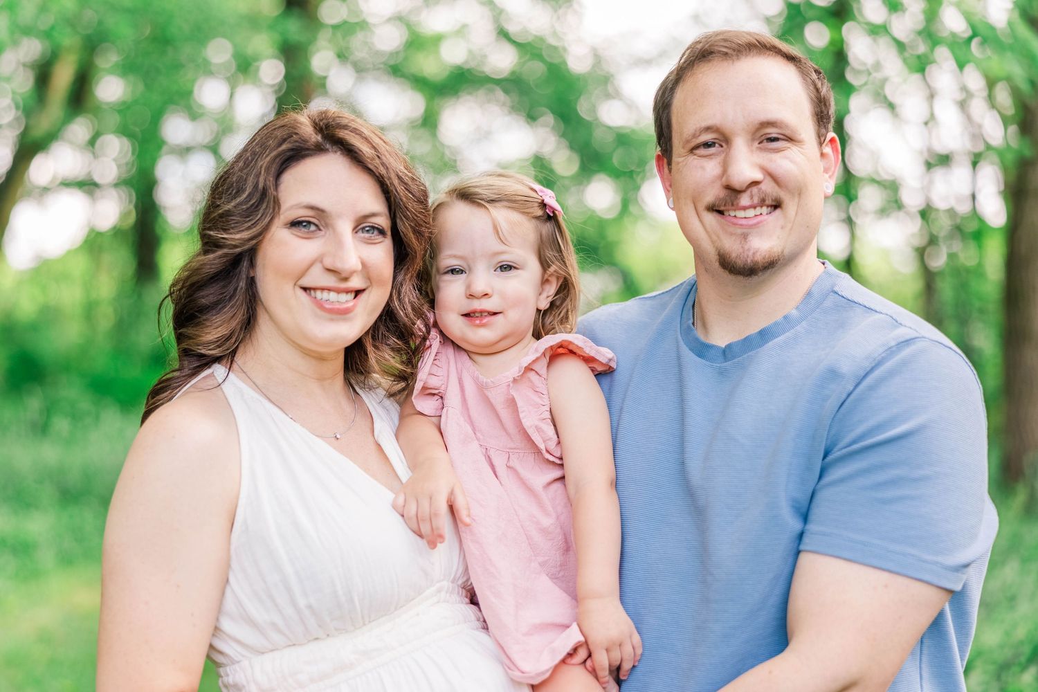 A young family poses for a portrait together outdoors with lush greenery in the background.