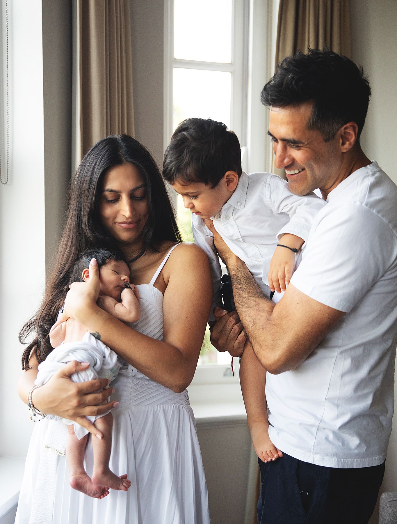 A family of four shares a tender moment together by a window in their home wearing matching white outfits. During a home newborn session