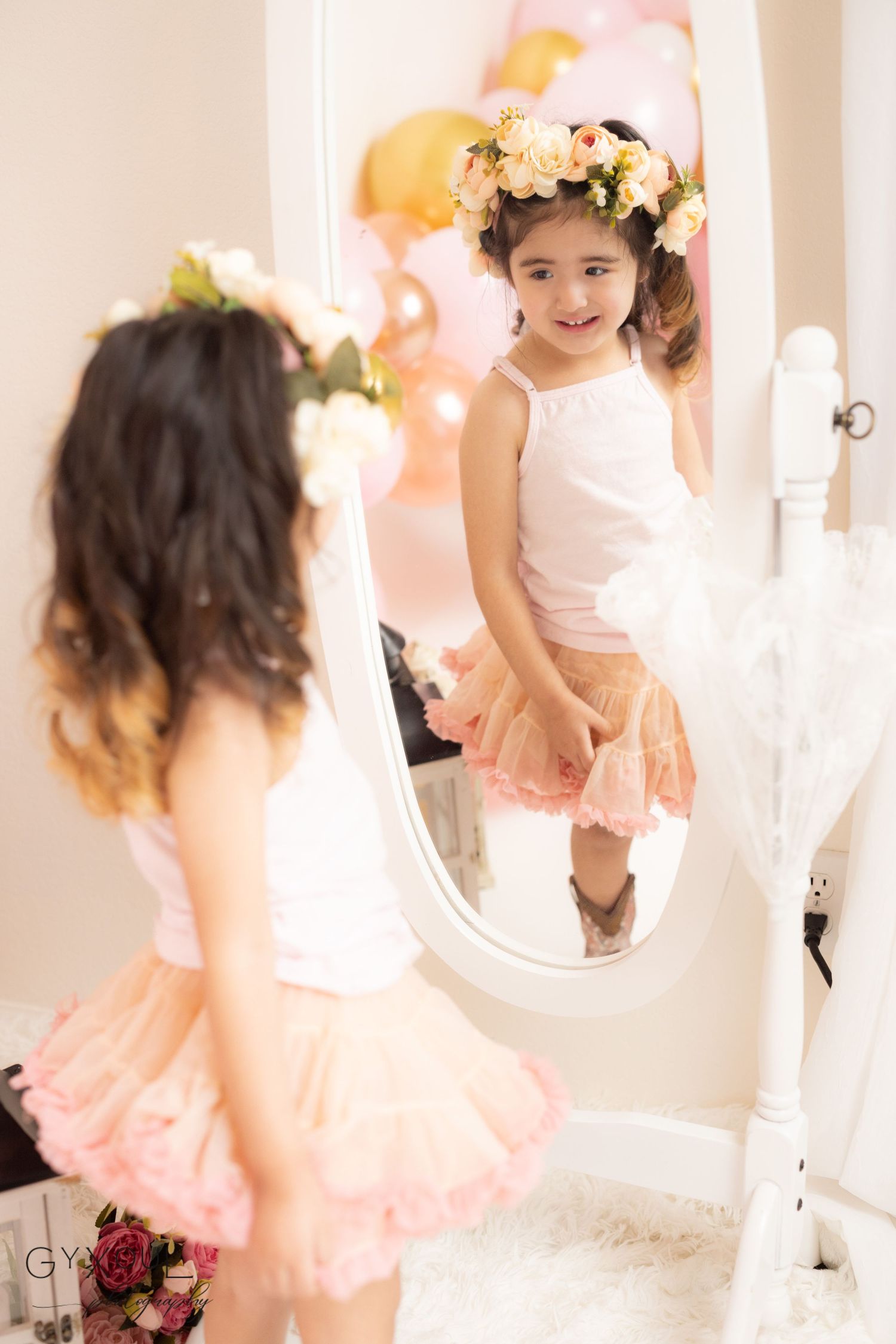 A child in a pink tulle skirt and floral crown poses in front of an ornate white mirror with soft pink lighting.