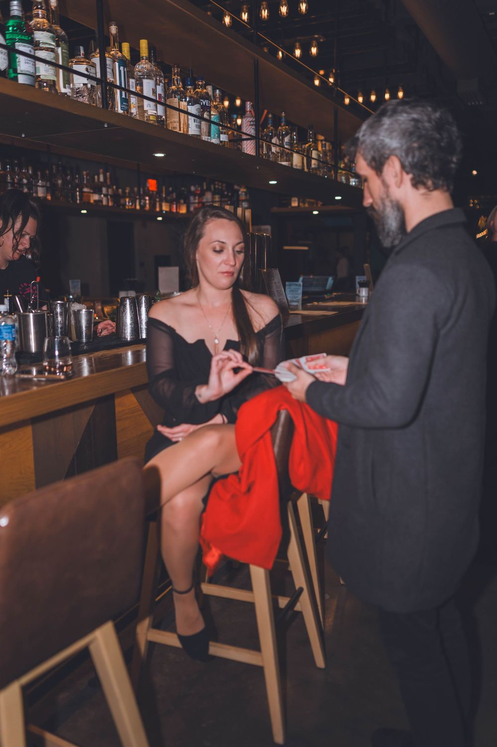 Bar patrons interact in an upscale cocktail lounge with bottles displayed on shelves and warm ambient lighting.