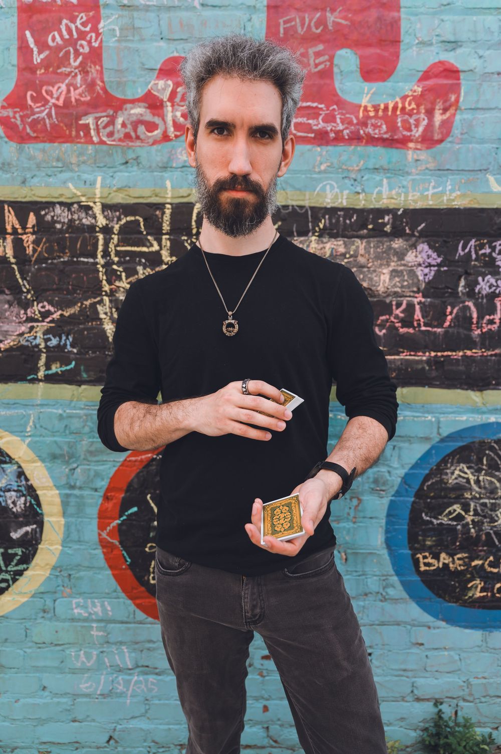 Person in black sweater stands against graffiti wall showcasing jewelry and tattoos.