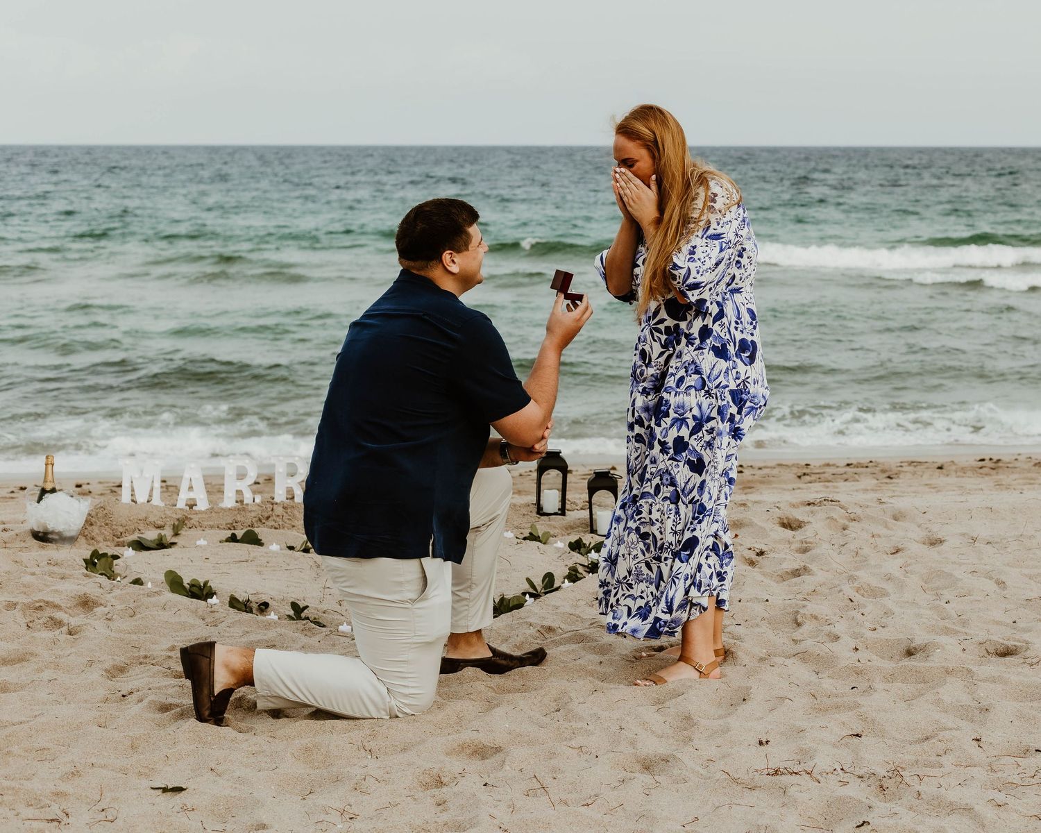 A couple sits together on a sandy beach with ocean waves in the background.