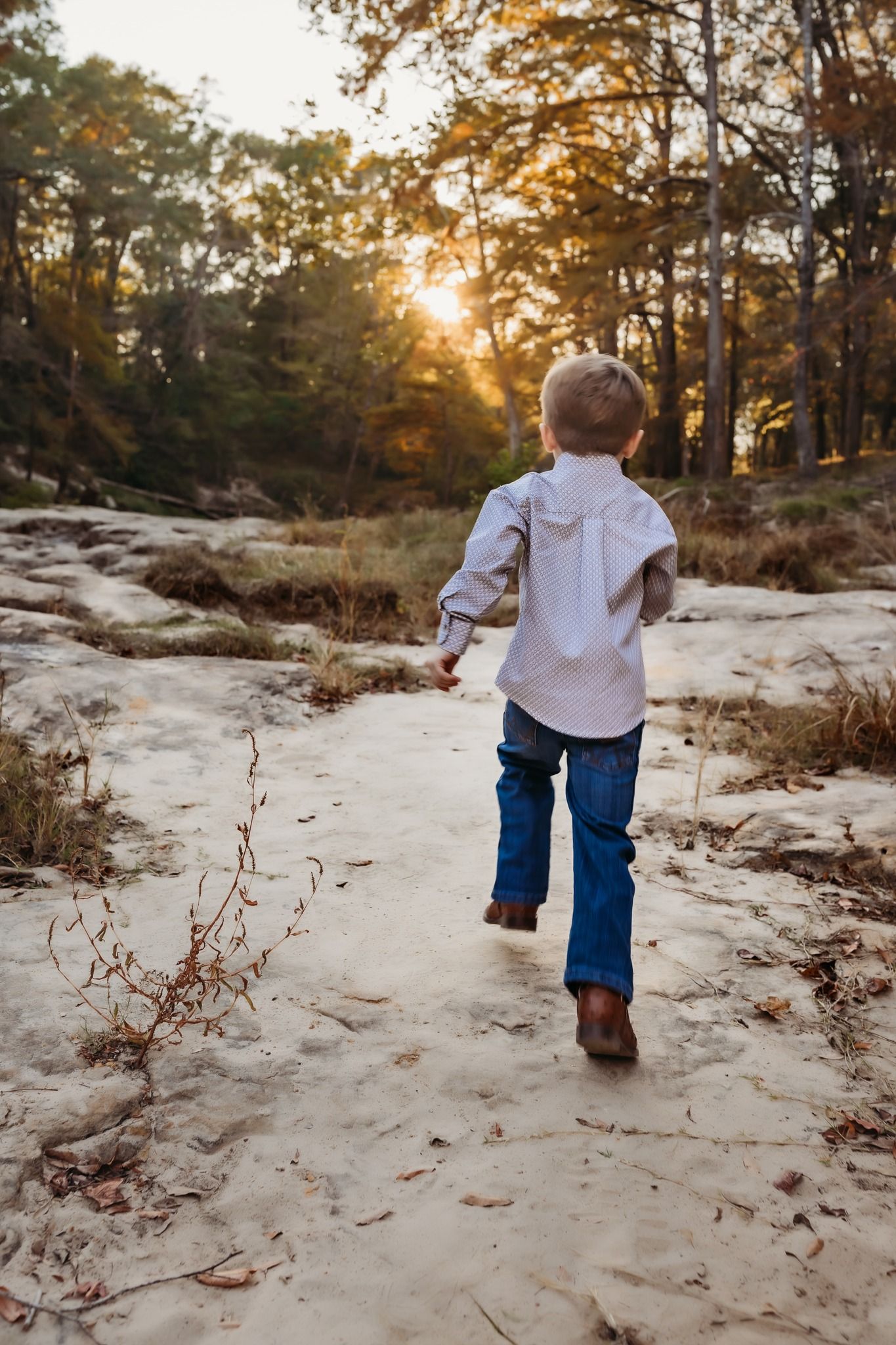 A small child runs along a sandy path through autumn woods at sunset, wearing jeans and a white shirt.