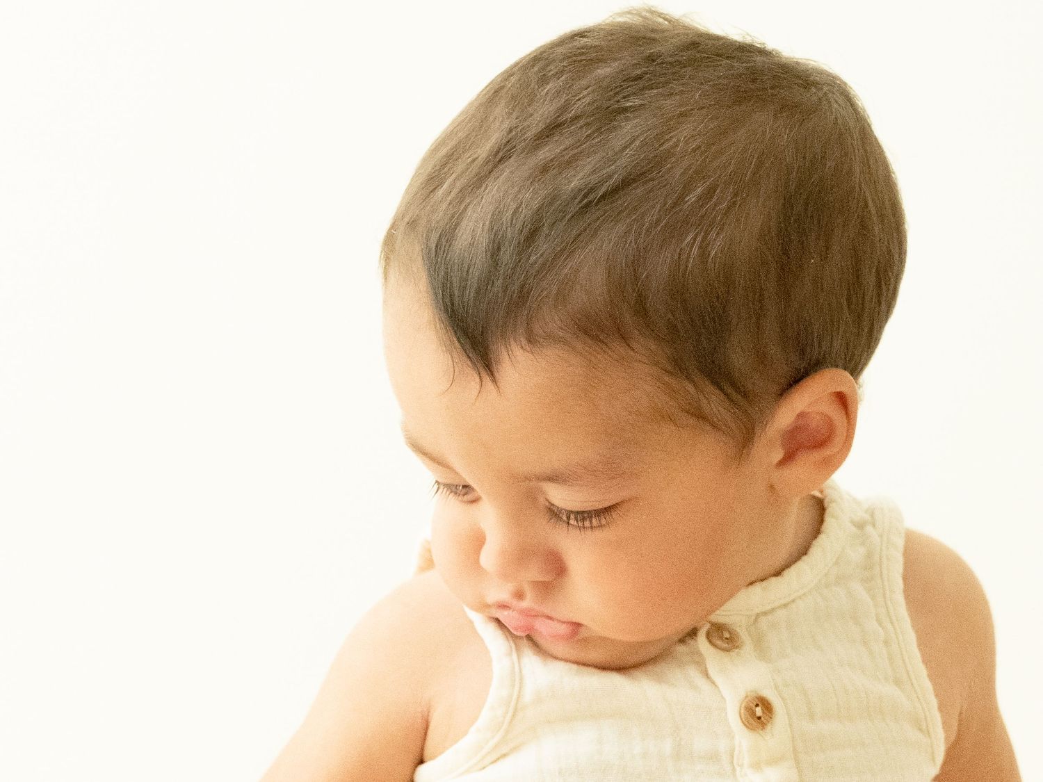 Small child wearing a light yellow sleeveless top looks down pensively against a white background.
