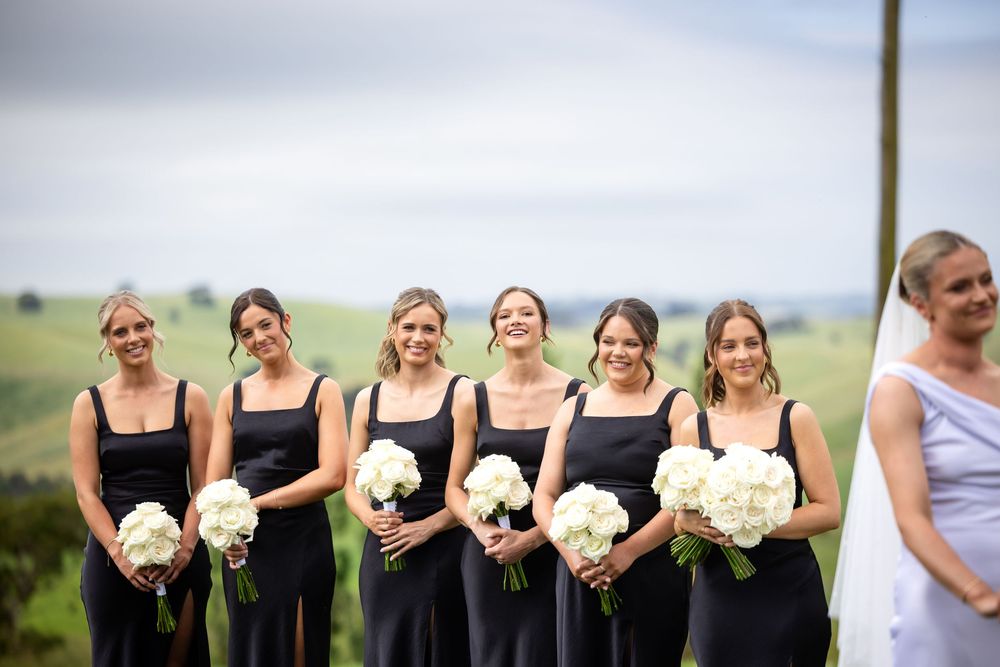 Wedding party members hold white floral bouquets in a line against a scenic outdoor backdrop.