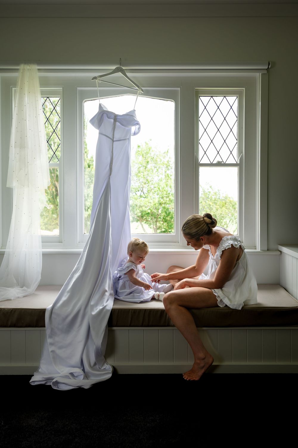 A tender moment by a window with sheer curtains and a flowing white dress hanging above on a sunny day.