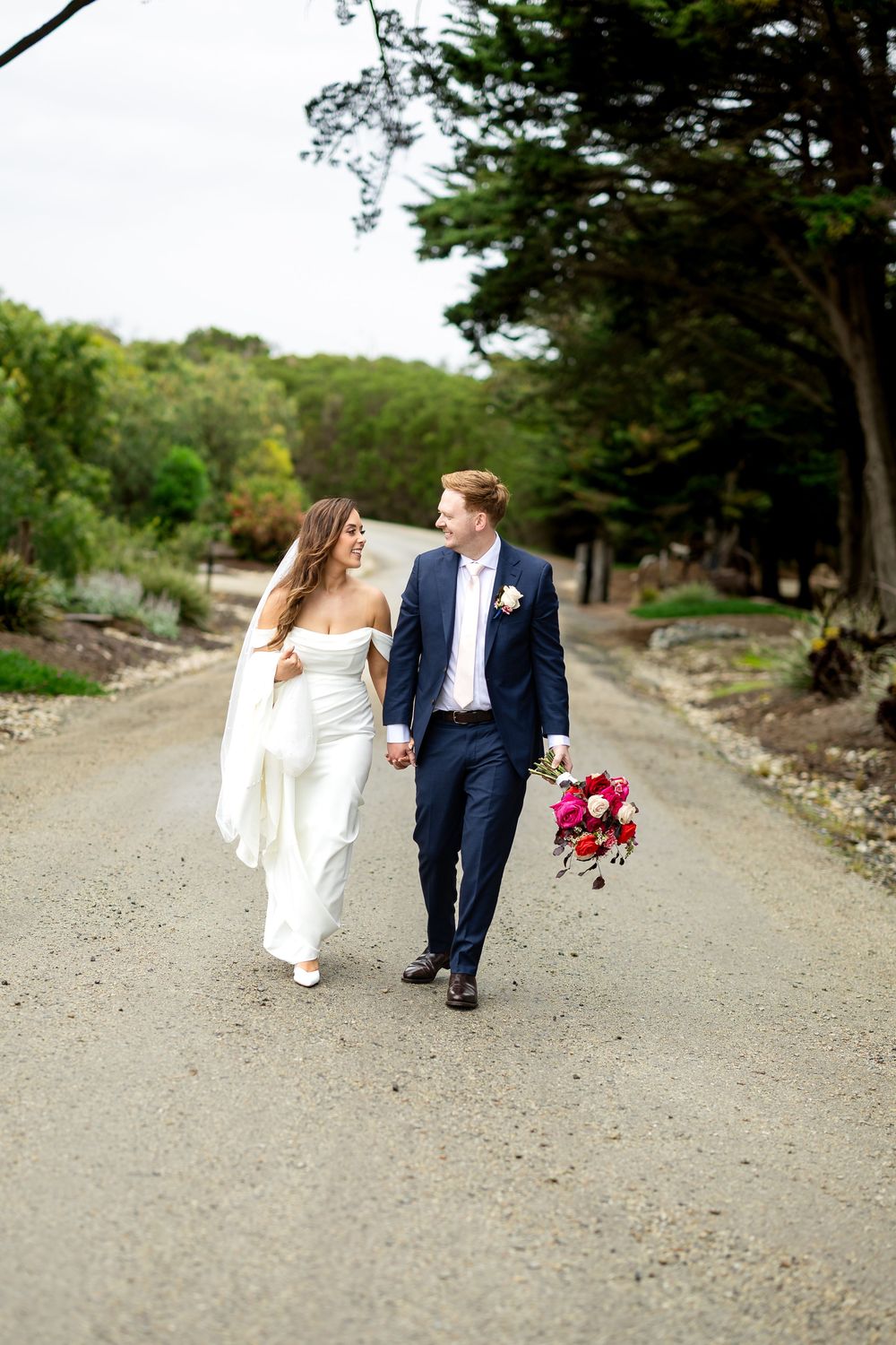 Bride and groom walk hand-in-hand down a tree-lined gravel path, bride in white gown holding vibrant pink bouquet.