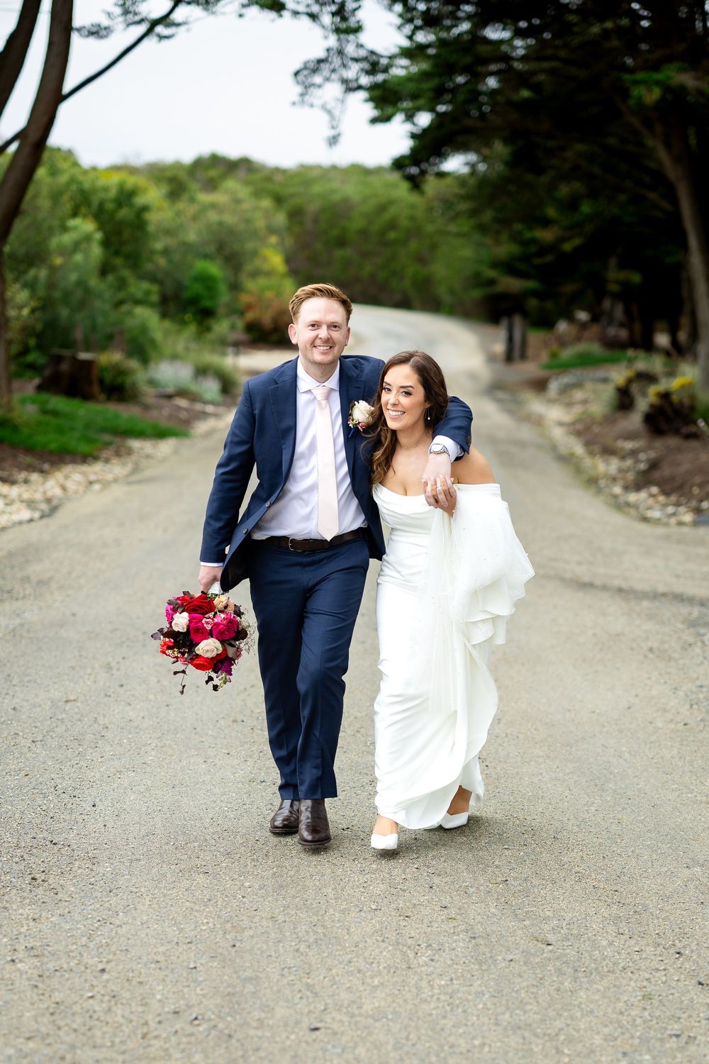 A happy couple walks down a tree-lined gravel path, bride in white gown, groom in navy suit holding red bouquet.