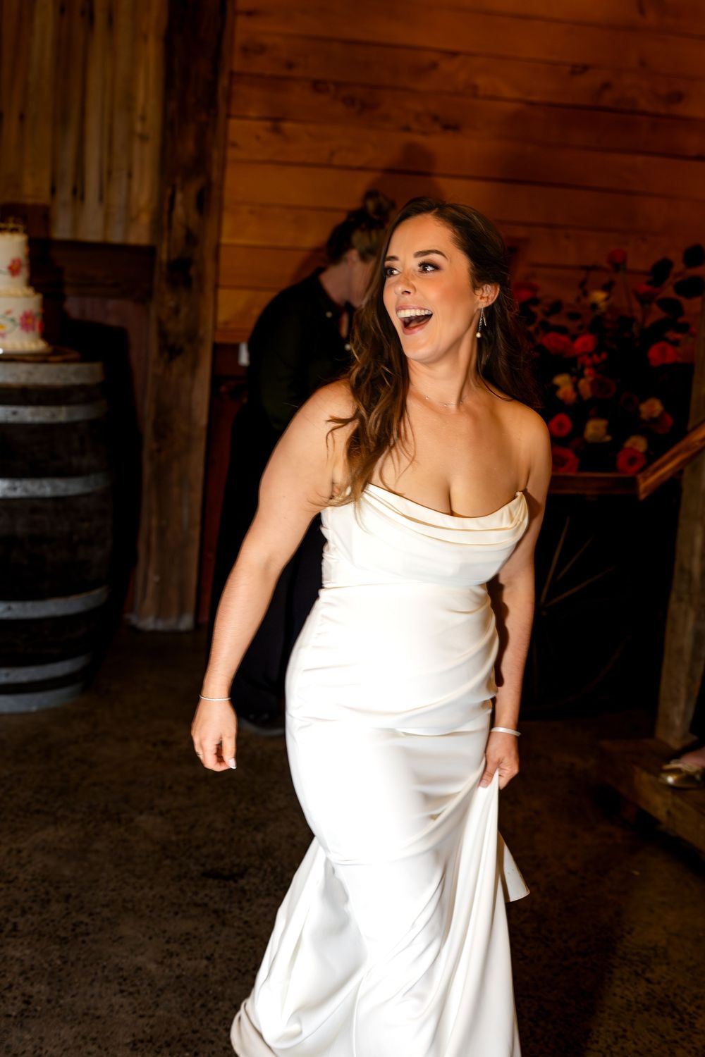 Bride in white satin gown laughing joyfully at rustic barn wedding reception with cake and floral arrangements.