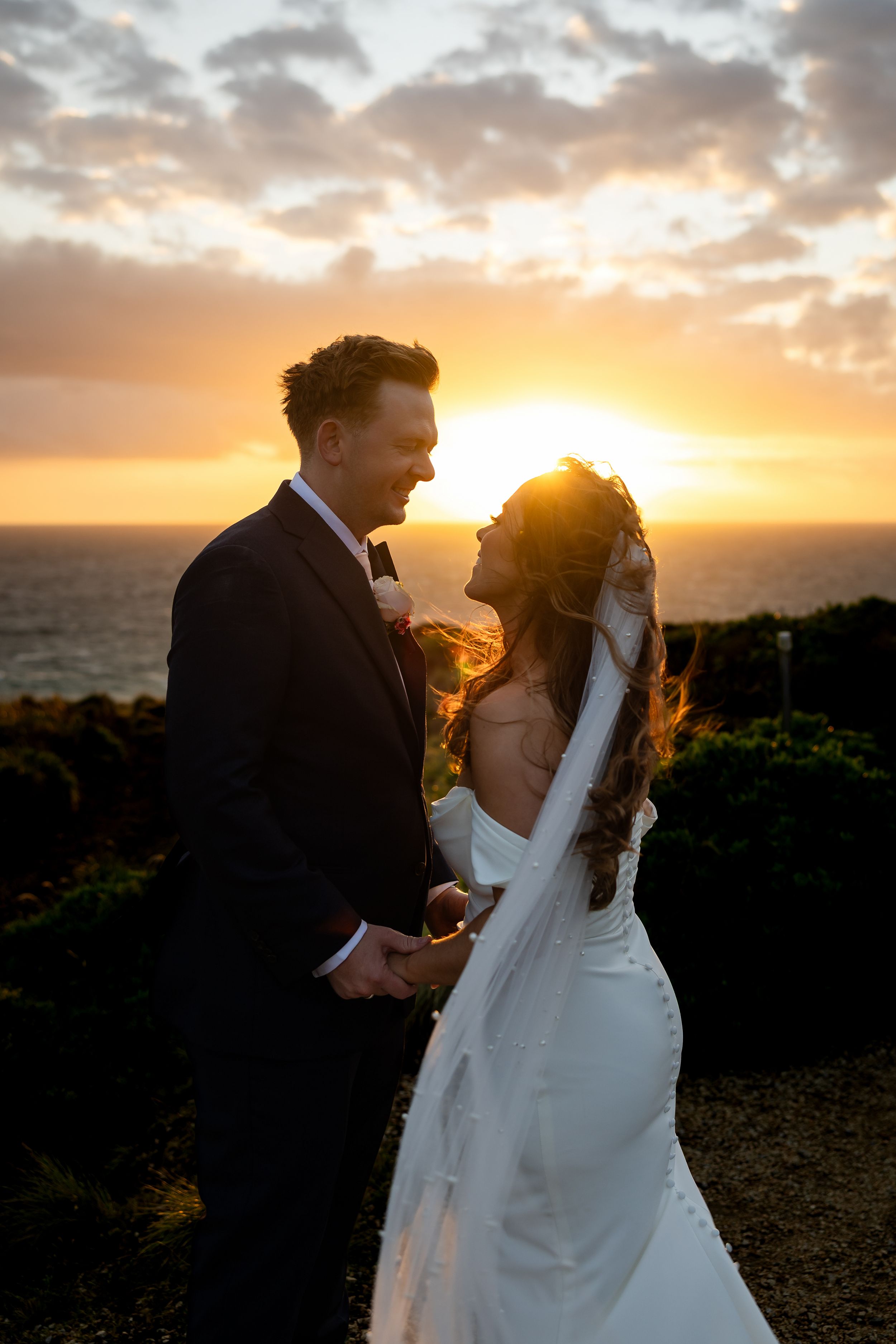 Bride and groom share a romantic moment at sunset by the ocean, golden light glowing behind them.