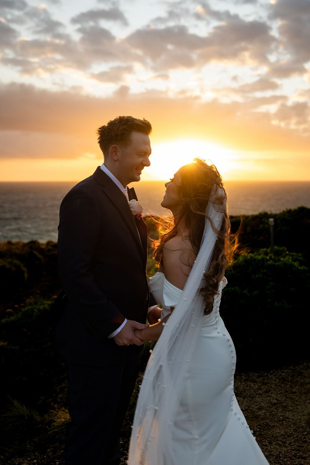 Bride and groom share a romantic moment at sunset by the ocean, golden light glowing behind them.