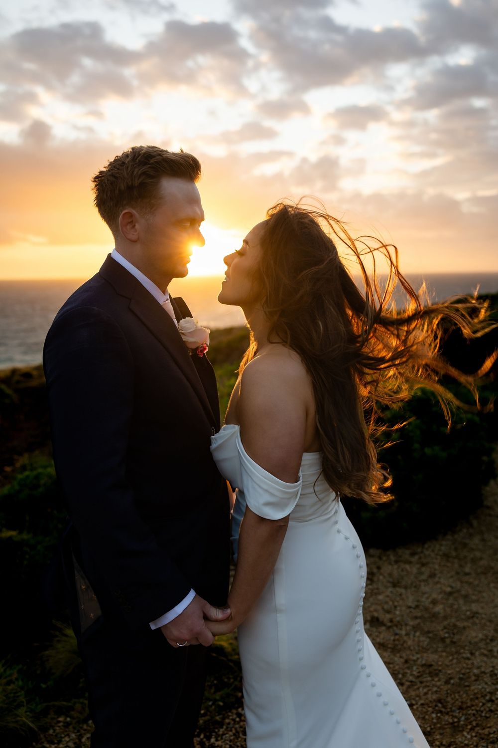 Bride and groom share a romantic sunset moment by the ocean, her hair flowing in the wind, wearing a white off-shoulder gown.