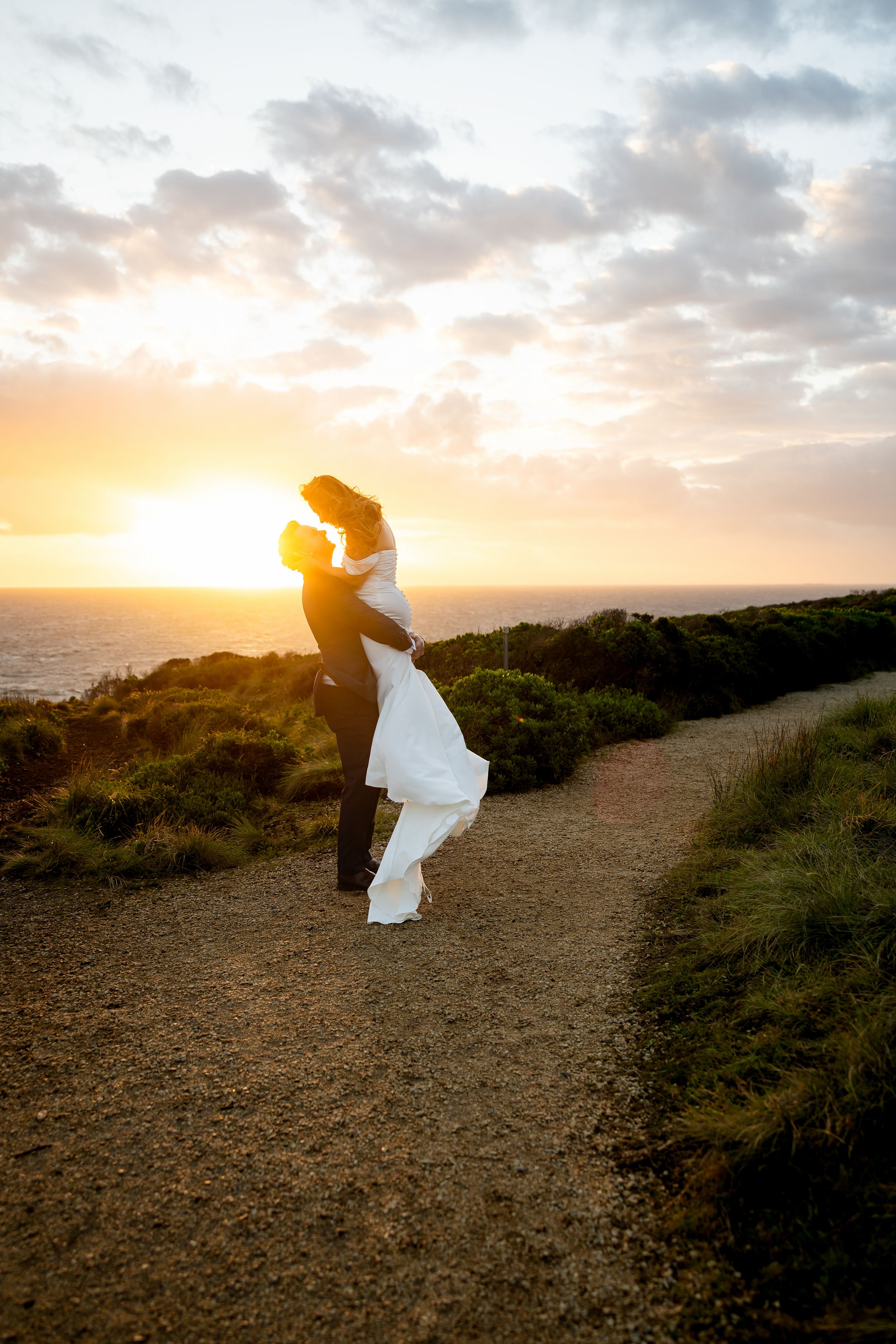 Couple embracing at sunset on coastal path, groom lifting bride in white dress with ocean and golden sky backdrop.