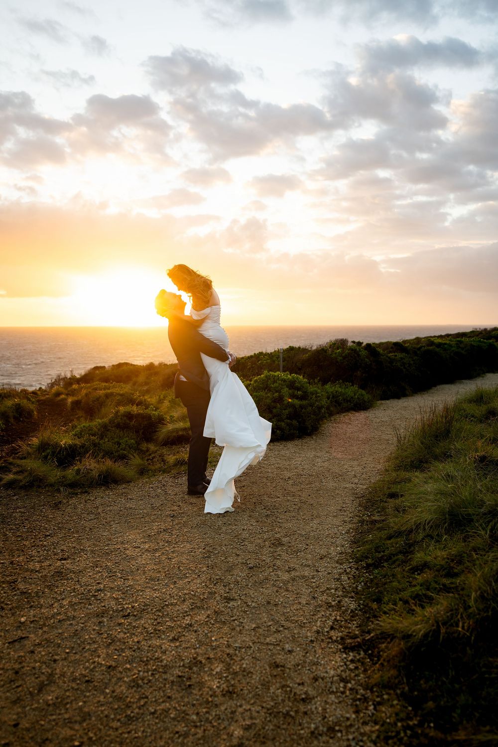 Couple embracing at sunset on coastal path, groom lifting bride in white dress with ocean and golden sky backdrop.