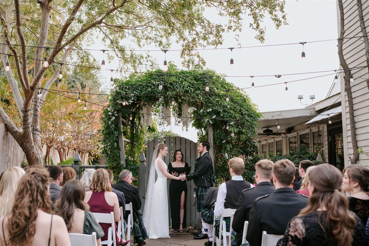 An outdoor wedding ceremony under a lush green floral arch with string lights and seated guests in a courtyard garden setting.