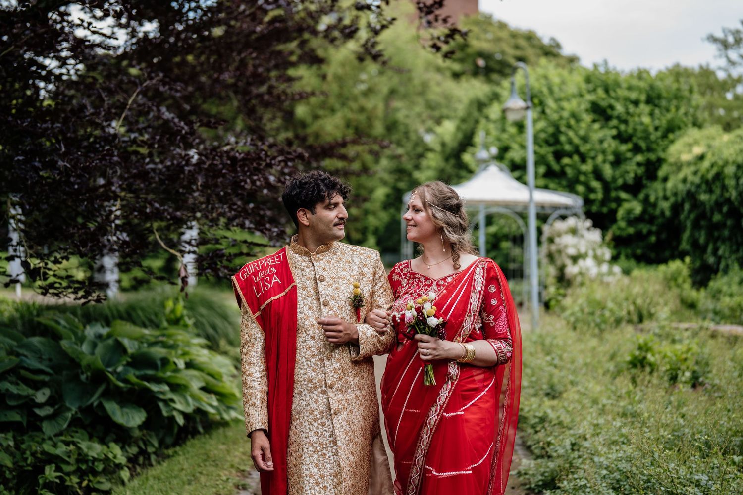 Couple in traditional red and gold wedding attire pose in lush garden setting.