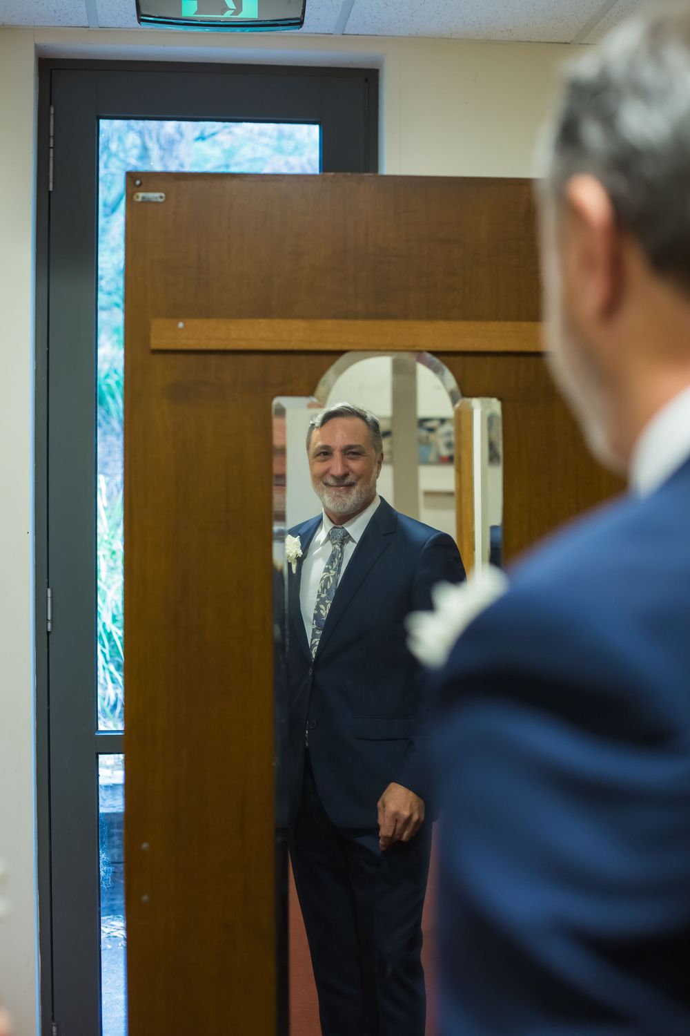 A well-dressed person in a navy suit looks at their reflection in a mirror within an office setting.