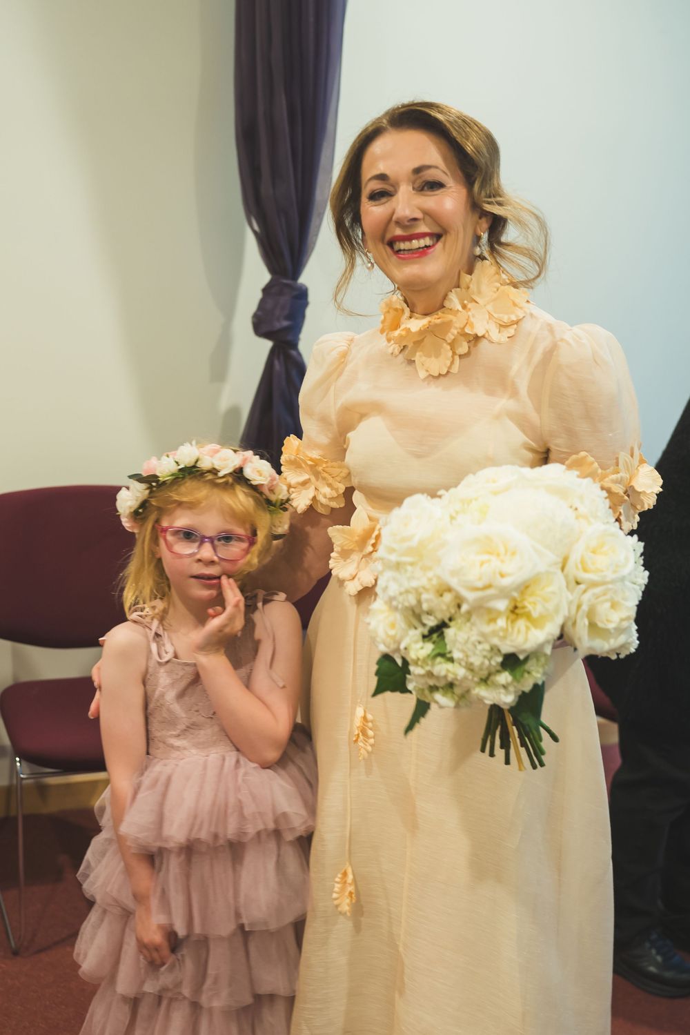 Two people share a joyful moment in formal attire while holding a bouquet of white flowers at an indoor venue.