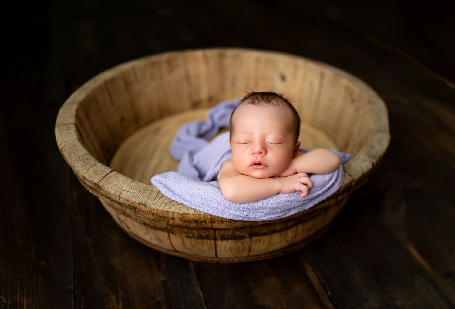 A small doll figure sits inside a curved wooden vintage dough bowl on a rustic wooden table surface.