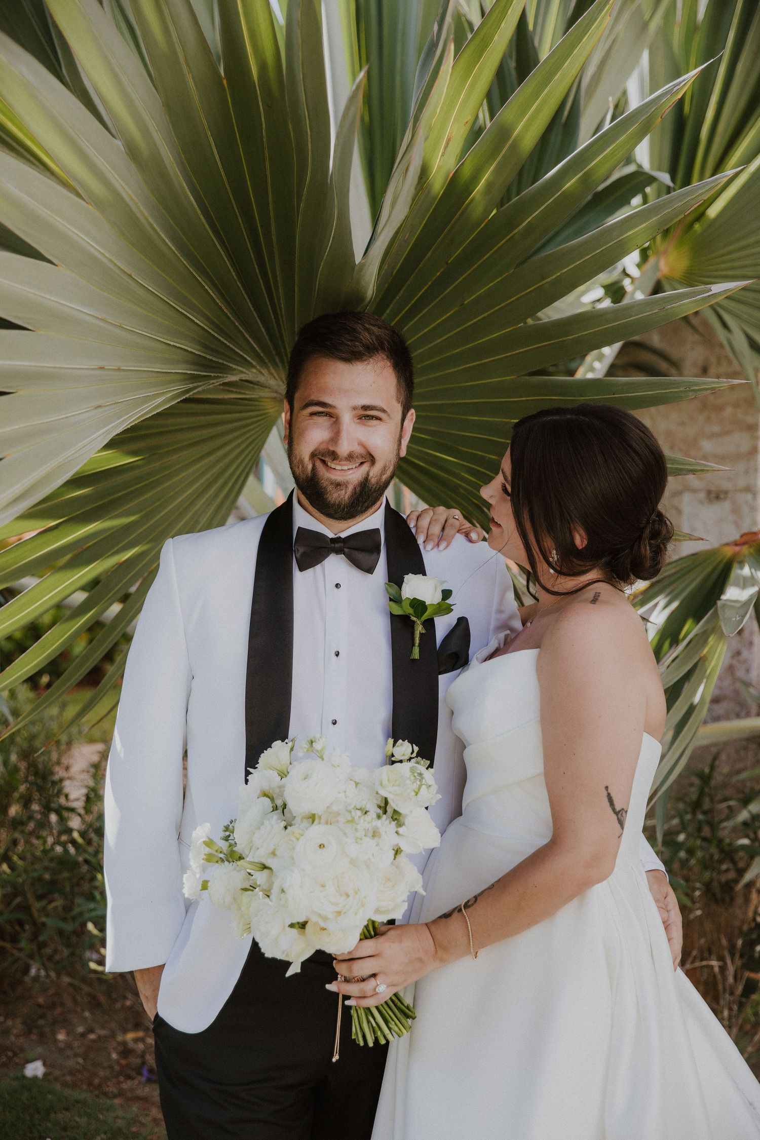 A wedding couple poses against tropical palm fronds with a white bouquet and formal attire.