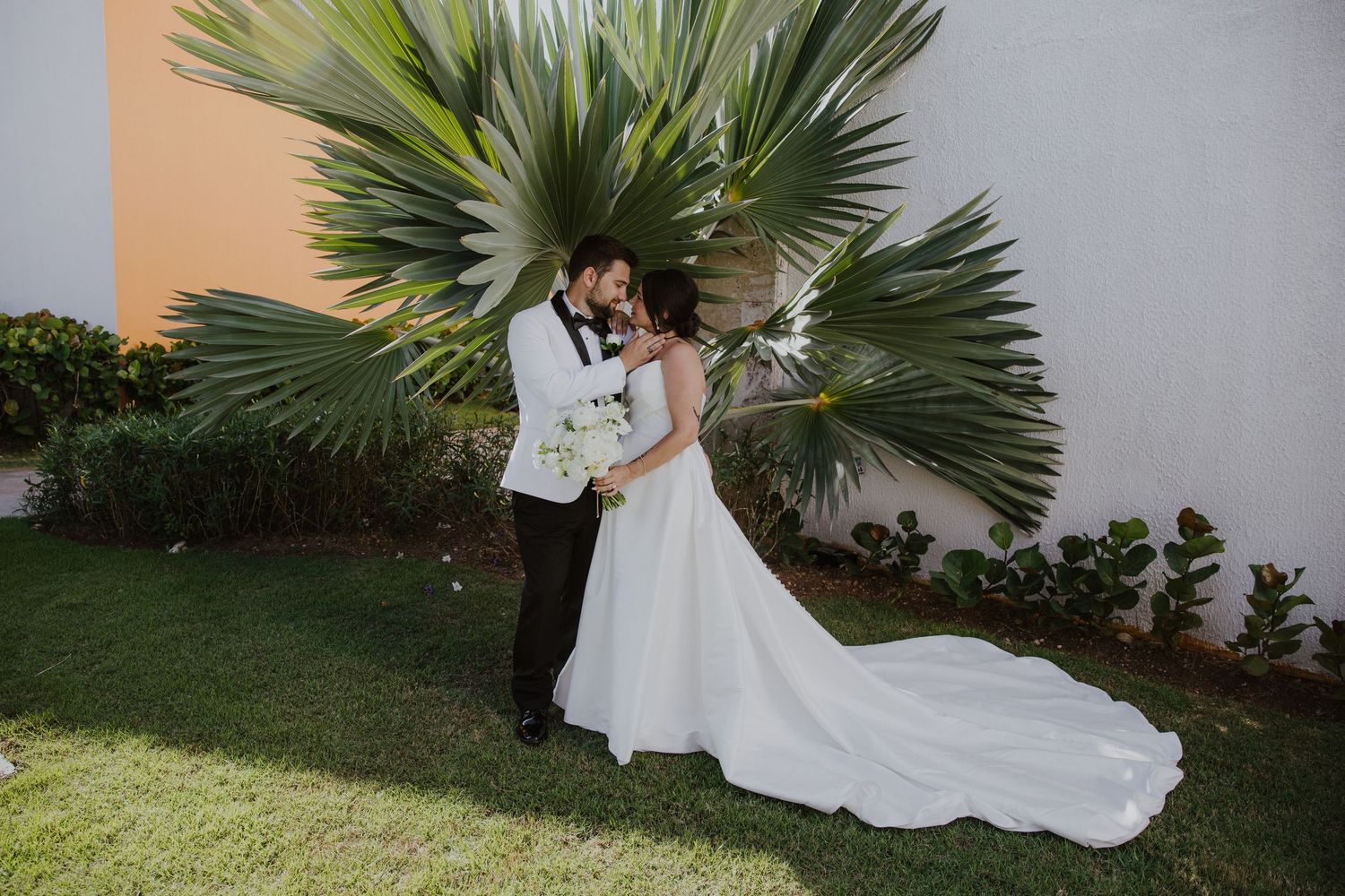 Newlyweds share an intimate moment under large palm leaves in a garden setting at Dreams Macao Beach resort