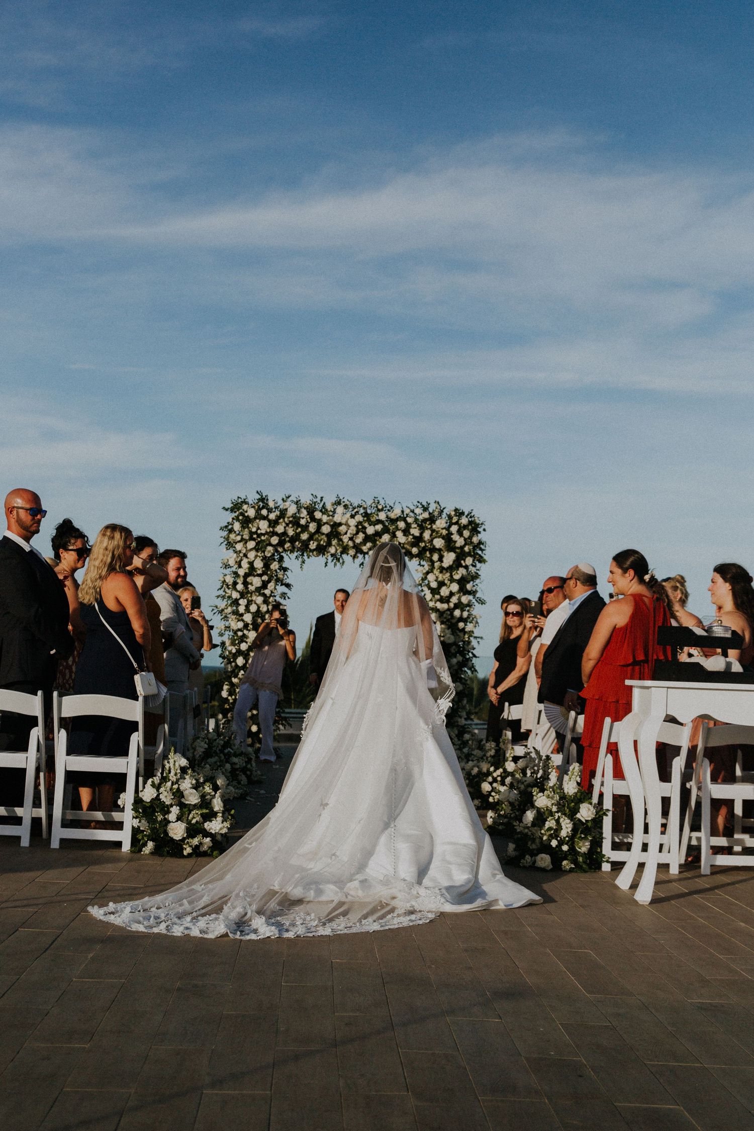 Wedding ceremony captured from behind showing bride's detailed lace train against floral arch backdrop.