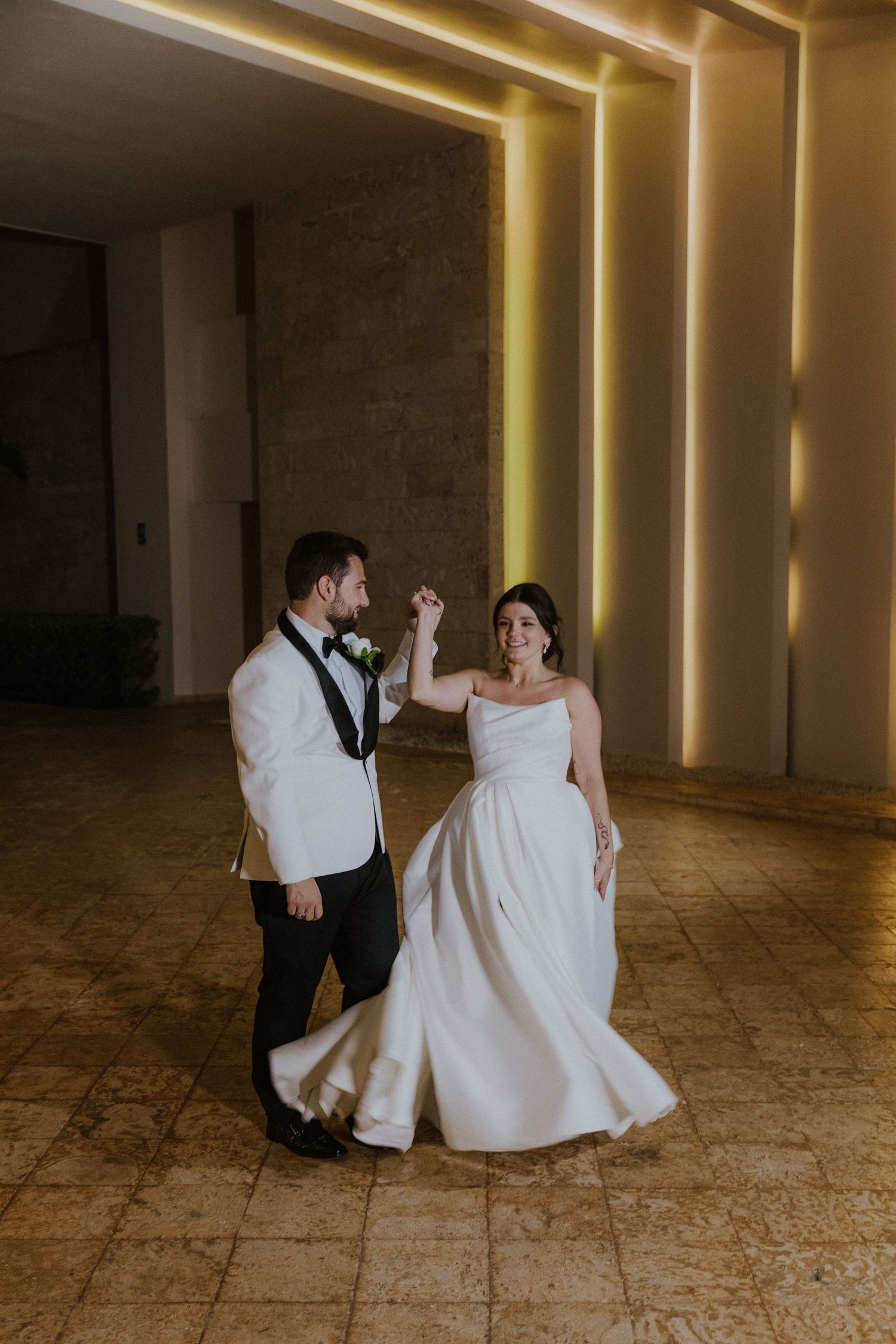 Elegantly dressed couple dancing on marble floor with backlit geometric wall patterns.