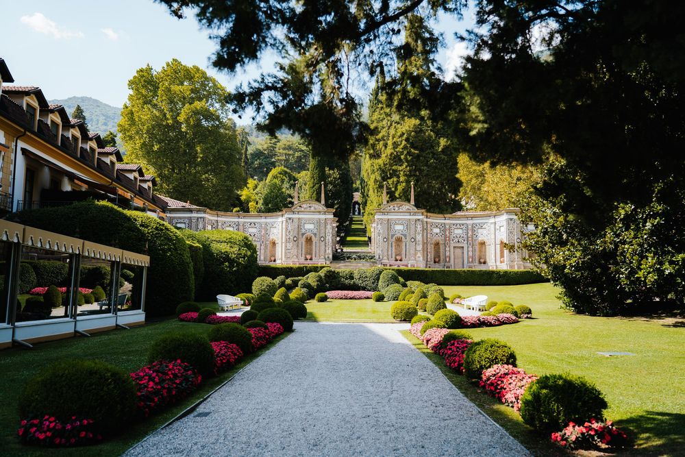 Red azalea bushes line a winding garden pathway through manicured grounds and historic buildings.