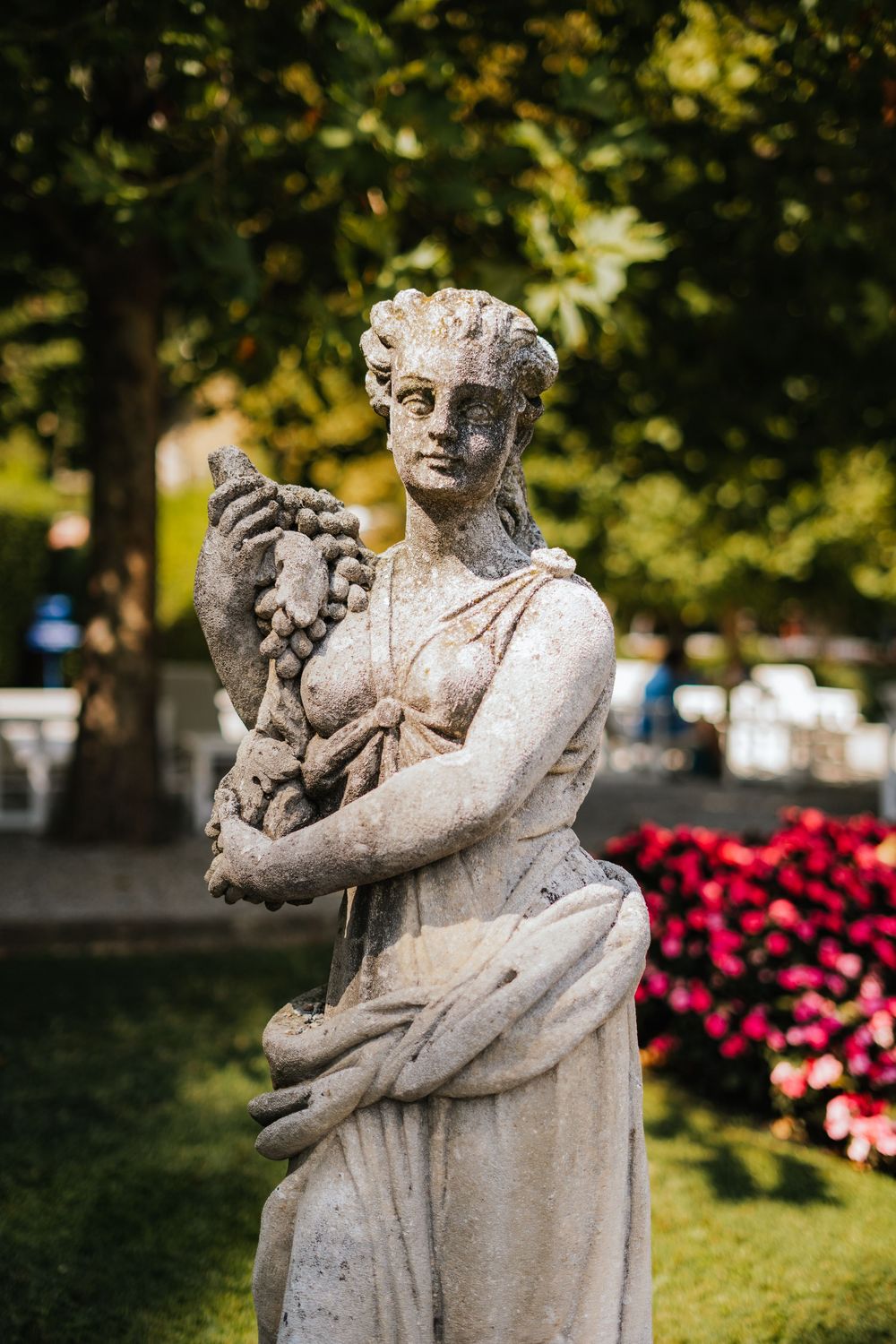 Stone sculpture of a classical-style female figure holding a cherub set in a garden with pink flowers and greenery.