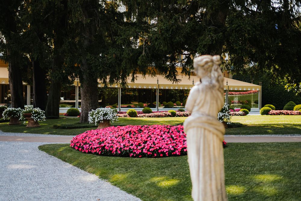 Classical white statue stands in a garden with vibrant pink flowers and manicured lawns at sunset.