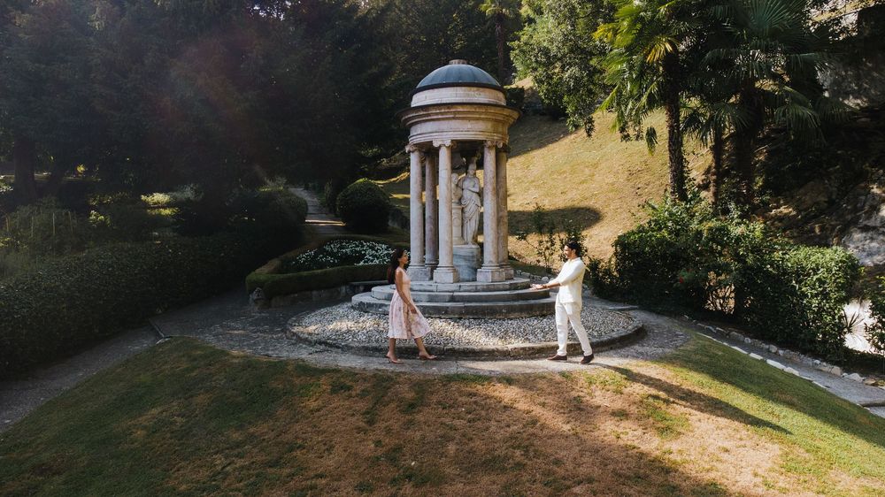 Two people stand near a classical stone gazebo with columns in a serene garden setting during golden hour.