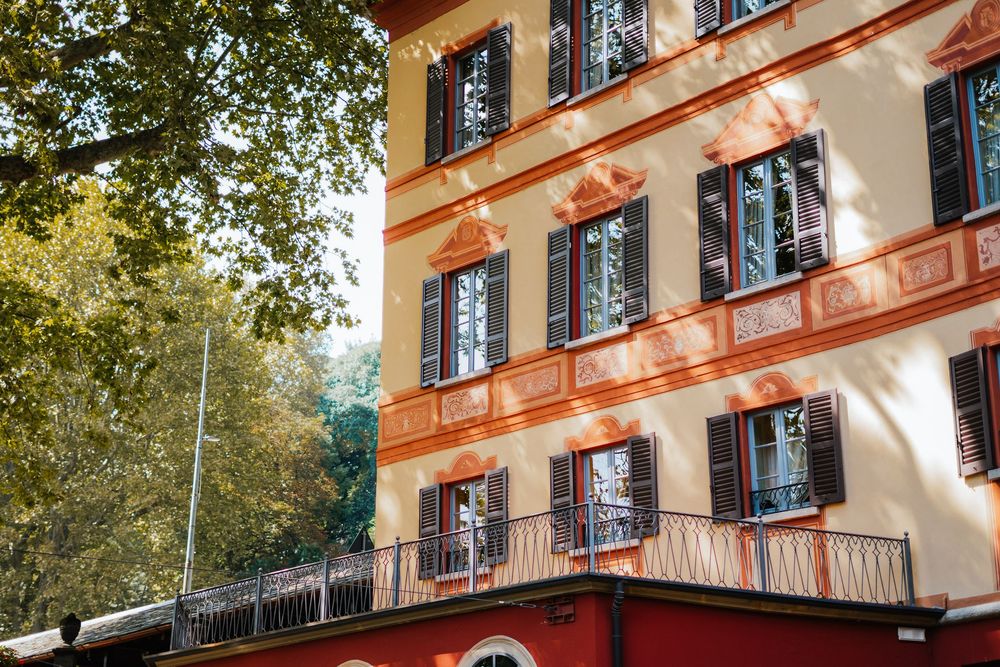 Orange painted European building facade with black shuttered windows and balcony railings against green trees on a sunny day.