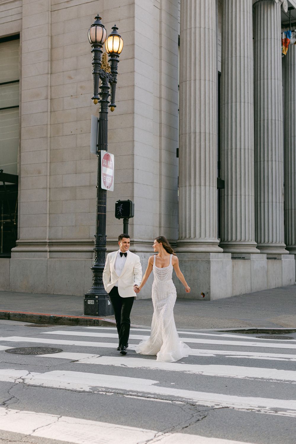 Bride in a white dress dances joyfully across a crosswalk in front of a grand building with columns.