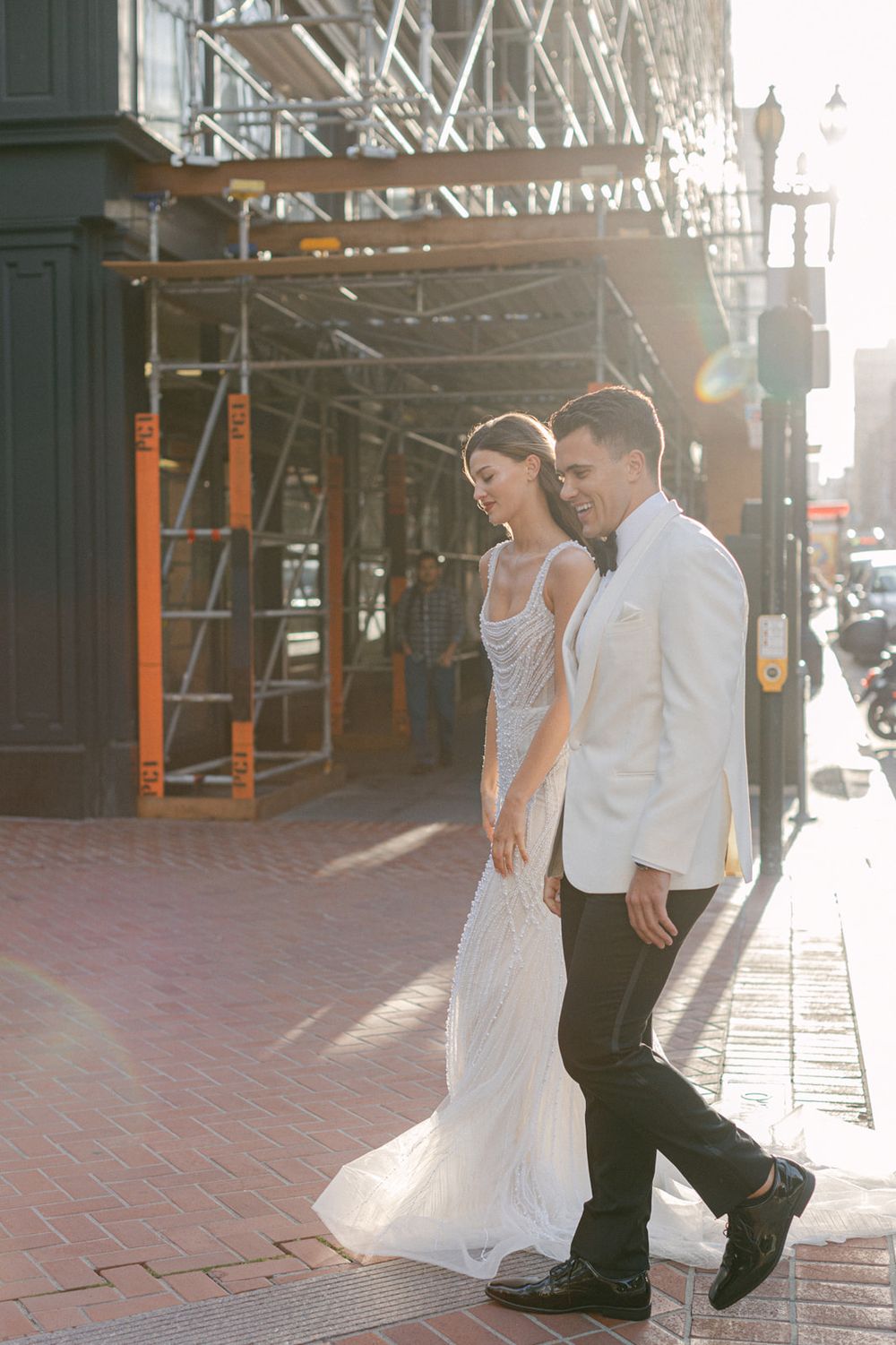 A couple walks together on a brick path in urban area surrounded by overhead scaffolding and architectural elements.