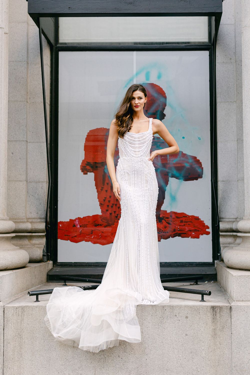 Elegant white mermaid wedding dress with beaded details and flowing train against abstract art backdrop.