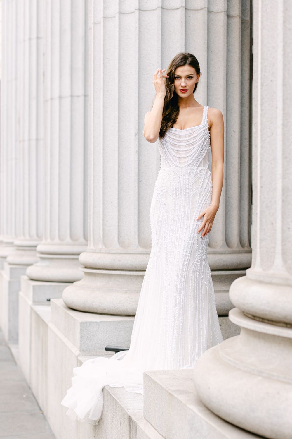 Elegant beaded white wedding gown with plunging neckline and curved silhouette photographed against classical stone columns.
