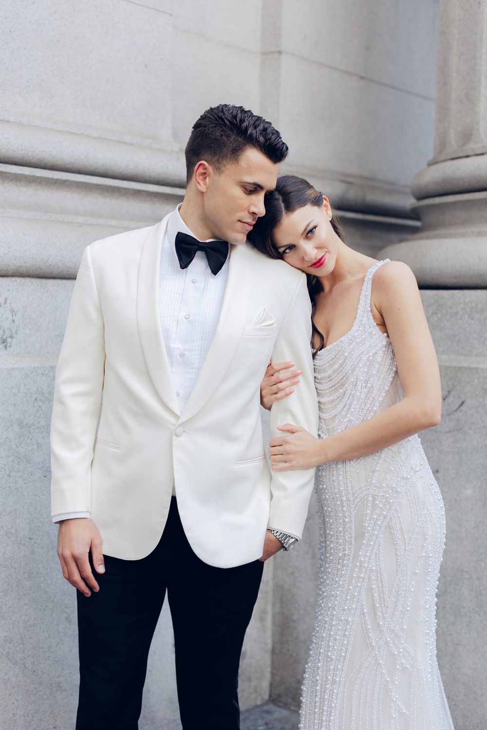 Elegant couple in formal attire pose against stone pillars, with white tuxedo jacket and beaded white gown.